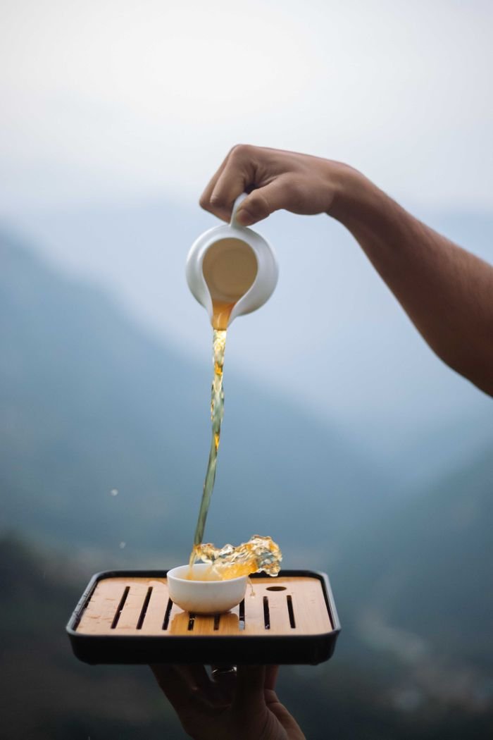 Hand gracefully pours hot tea onto a wooden tray outdoors.