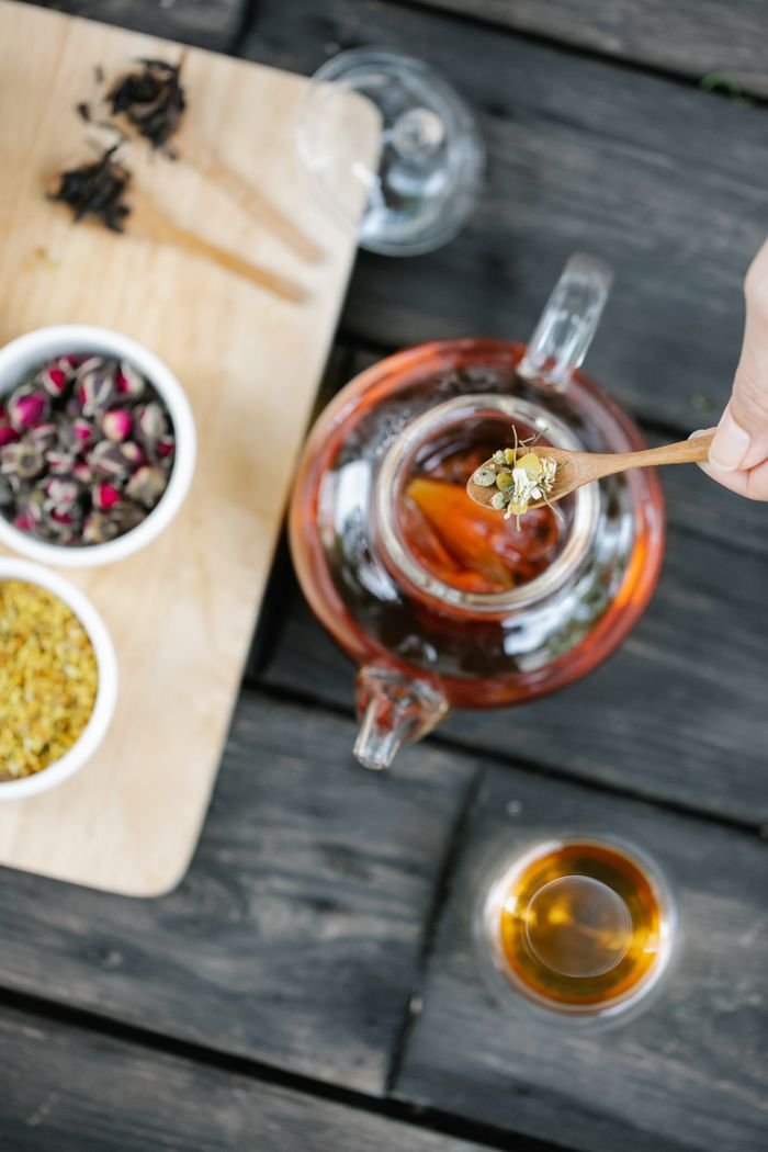 Overhead view of tea being prepared with assorted spices and flowers on a wooden tray.