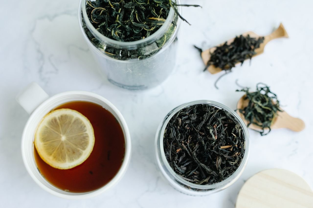 Close-up shot of a cup of tea with a lemon slice and jars of loose leaf tea on a marble surface.